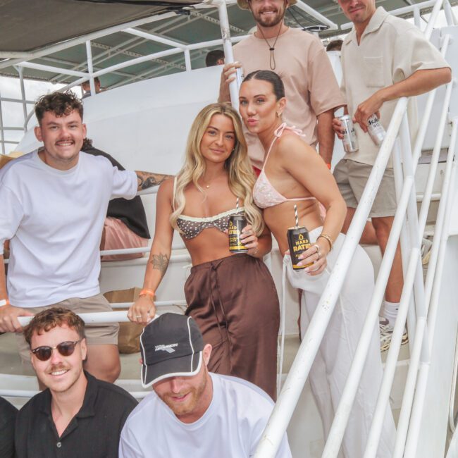 A group of eight young adults pose and smile on the stairs of a boat. Some hold drinks and wear casual summer outfits. The setting appears festive and relaxed, with bright daylight and outdoor seating visible.