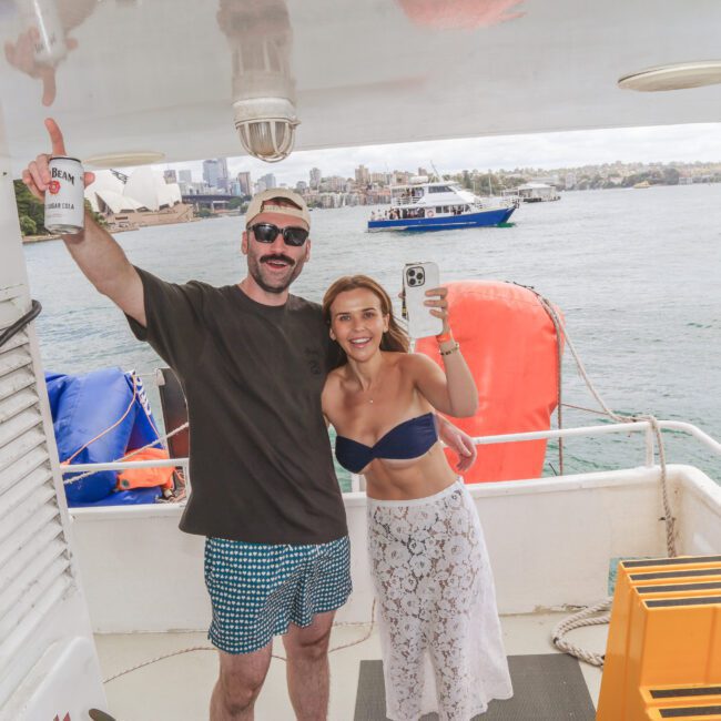 A man and woman smiling and posing for a selfie on a boat deck, with water, boats, and a city skyline visible in the background. The man raises a drink, and both appear to be enjoying a sunny day.