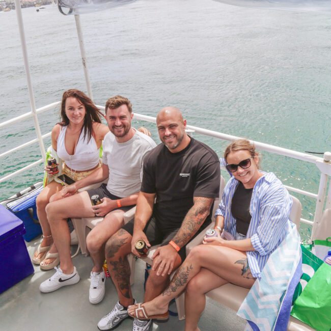 Four people sit side by side on a boat, smiling at the camera. Two women and two men appear relaxed, holding drinks, with water in the background and bags on the deck near them.