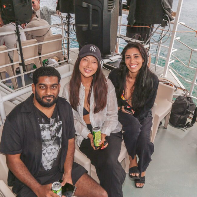 Three people sit and smile on a boat deck, each holding a can of drink. Behind them, speakers and other passengers are visible. The ocean can be seen through the boat railings.