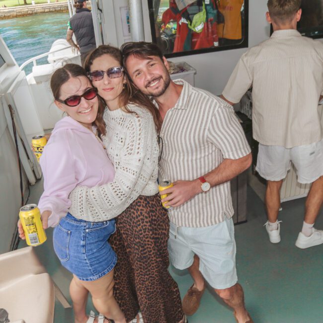 Three people smile and pose together on a boat, each holding a can of drink. They wear casual summer clothes and sunglasses. Other people are in the background, and water is visible outside. “Yacht Social Club” logo is in the corner.