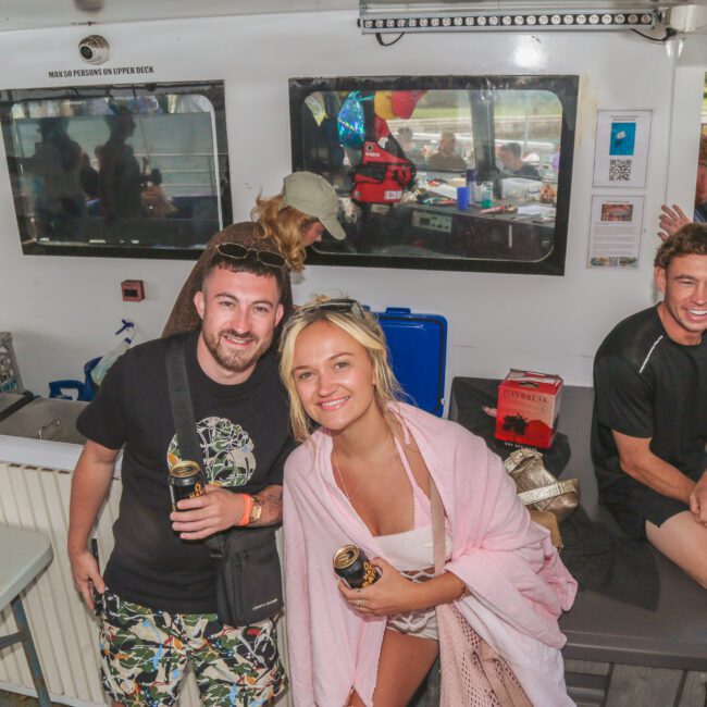 A group of people enjoy a boat party. Two people in the foreground smile at the camera, holding drinks, while others relax and chat in the background near windows and coolers. The atmosphere is casual and festive.