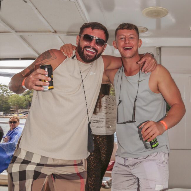 Two young men smiling with arms around each other, holding drinks, standing on a boat. One wears sunglasses and checked shorts, the other wears a tank top and shorts. People and water are visible in the background.
