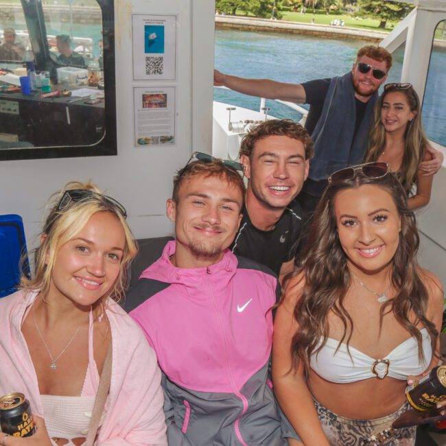 A group of five young adults smile and pose for a photo inside a boat, holding cans of drinks. Sunlight streams in through the windows, and a man waves from outside. Water and greenery are visible in the background.