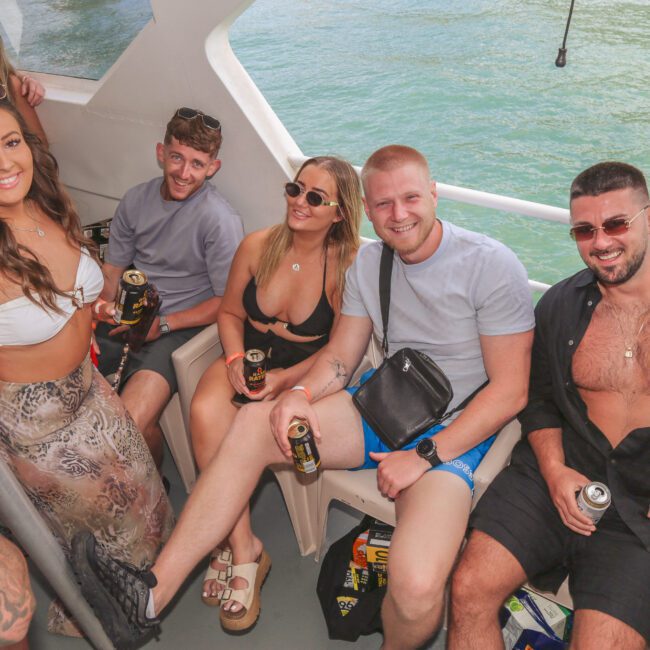 A group of five young adults smile and pose for a photo on a boat, holding drinks, with sunny weather and blue water in the background.