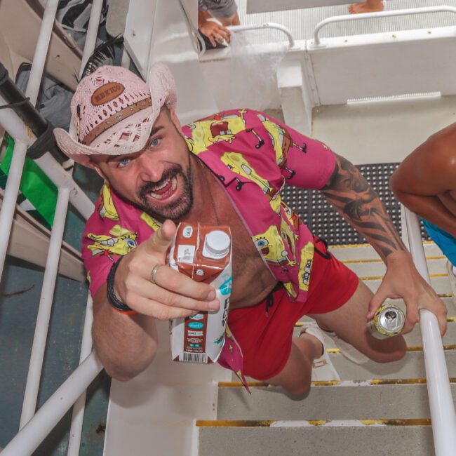 A man in a pink cowboy hat and colorful shirt excitedly poses on stairs with drinks in hand, next to a man in sunglasses and swim trunks. Both are on a boat, and the scene looks lively and festive.