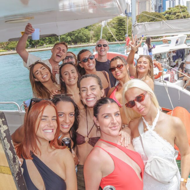 A group of people, mostly women in swimsuits and sunglasses, smile and pose on a yacht with water and city buildings in the background. Another yacht with people is seen nearby. It’s a sunny day.