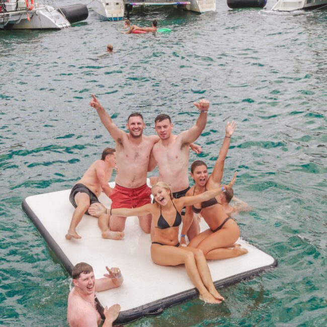 A group of young adults in swimsuits smile and pose on a floating dock in clear blue water, with boats and other people swimming in the background. Everyone looks happy and excited, enjoying a fun day together.