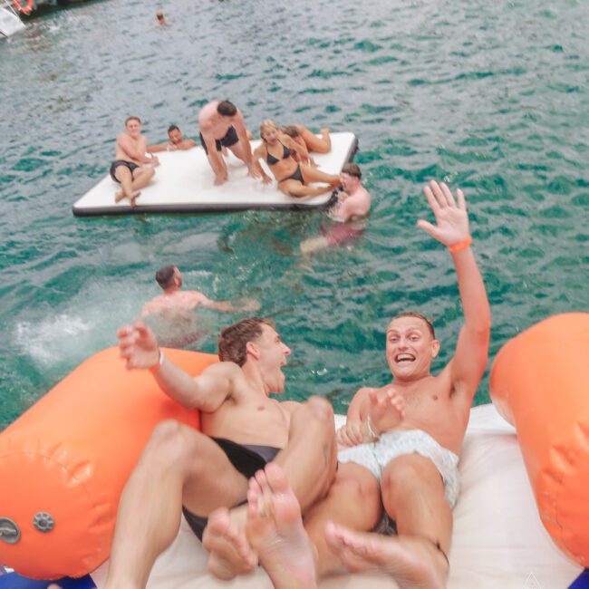 Two young men slide down an inflatable water slide into a lake, smiling and waving, while people relax and swim on a floating mat in the background. Boats are visible on the water.