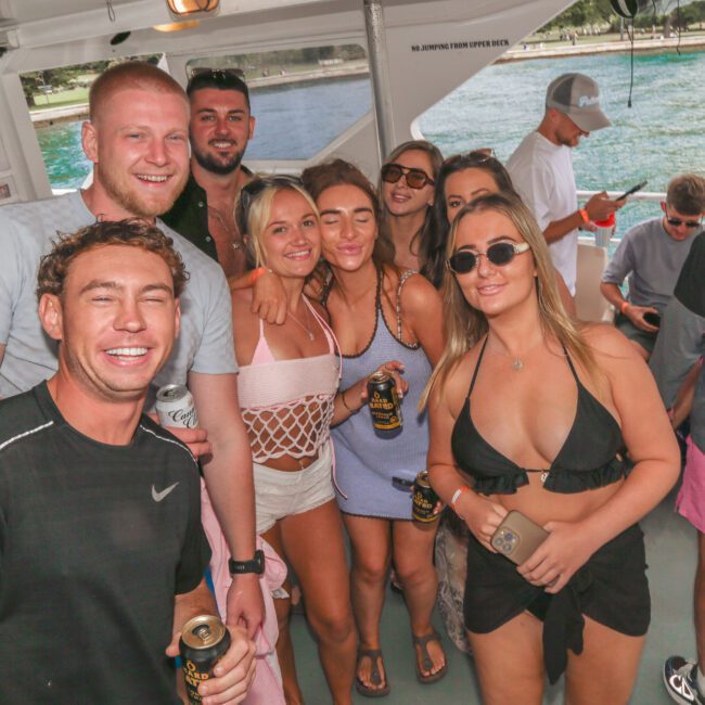 A group of young adults smiles and poses together on a boat, some holding drinks. The group is dressed in casual summer clothing and swimwear, with water and trees visible in the background.