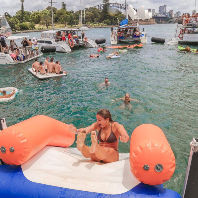 A woman in a bikini slides down an inflatable slide from a boat into the water, surrounded by people swimming and relaxing on boats and floats in a lively harbor scene. The Sydney Harbour Bridge is visible in the background.