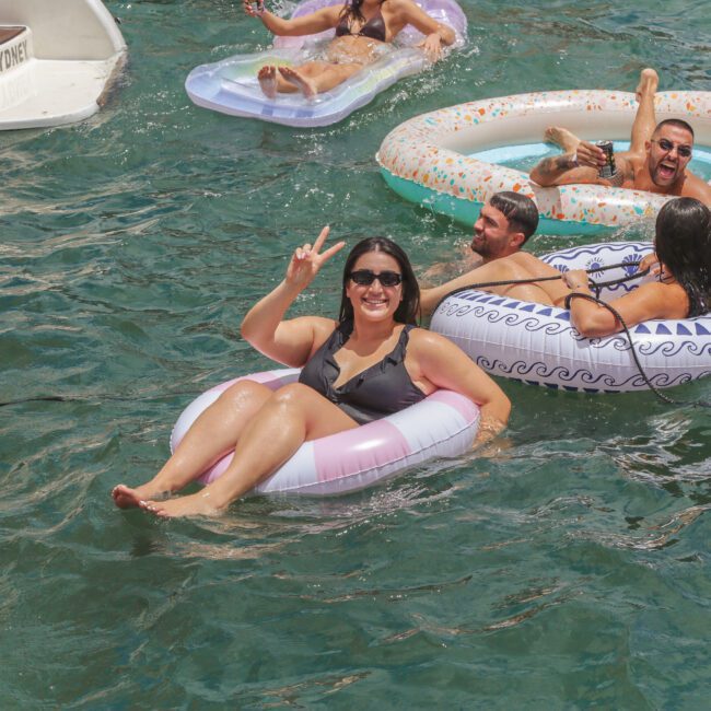 A woman in sunglasses smiles and flashes a peace sign while floating on a pink and white inflatable ring in the water, surrounded by others relaxing in colorful tubes. A boat is partially visible in the background.