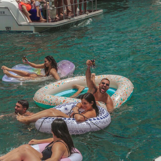 People relax on inflatable floats in clear blue water near a boat. Some are holding drinks and smiling, enjoying a sunny day with others nearby. A logo in the corner reads “White Social Club.”.