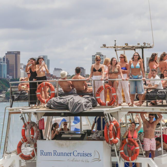 A group of people are dancing and socializing on the upper deck of a Rum Runner Cruises boat, with a city skyline and harbor in the background under a partly cloudy sky.