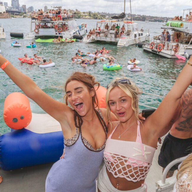 Two women smiling with arms raised on a boat, holding drinks, with people on floats and other boats in the water behind them, enjoying a lively summer party.