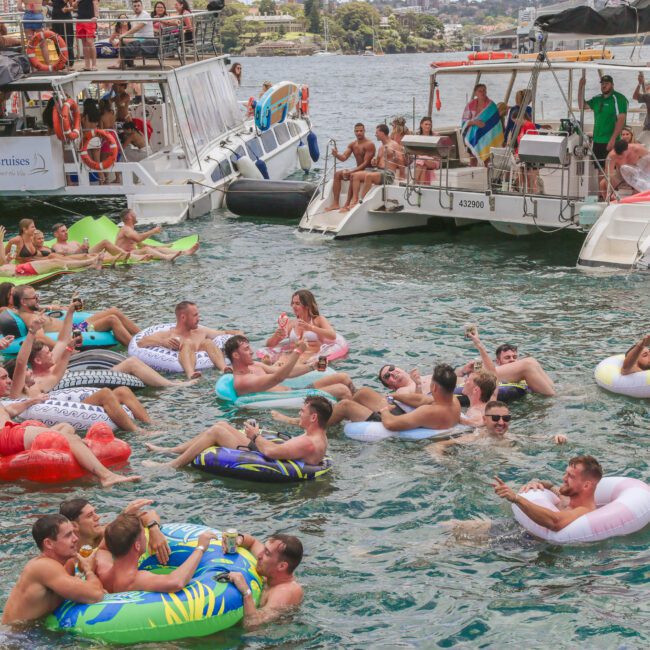 A group of people float on colorful inflatables in the water near two docked boats, enjoying a lively, sunny day. Some people are on the boats watching, while others relax and socialize in the water.