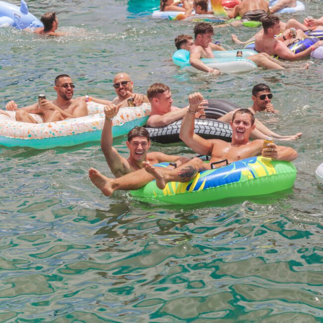 A group of people relax and have fun on colorful inflatable floats in a crowded body of water, waving, smiling, and enjoying a sunny day together. Some hold drinks; the atmosphere is lively and social.