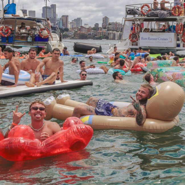 A group of people relax on colorful inflatables and boats in a busy harbor, with city buildings and a bridge in the background. Most are in swimsuits, enjoying the sunny day on the water.