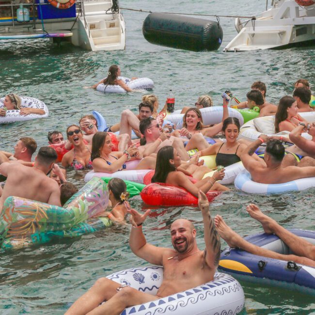 A large group of people relax and have fun on inflatable pool floats in the water near boats, enjoying a lively summer party atmosphere. Many are smiling, laughing, and raising their arms in celebration.