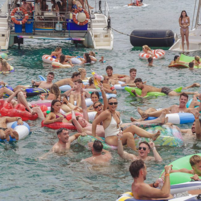 A large group of people are enjoying a party in the water, floating on colorful inflatables near anchored boats. They are smiling, talking, and having fun under sunny weather.
