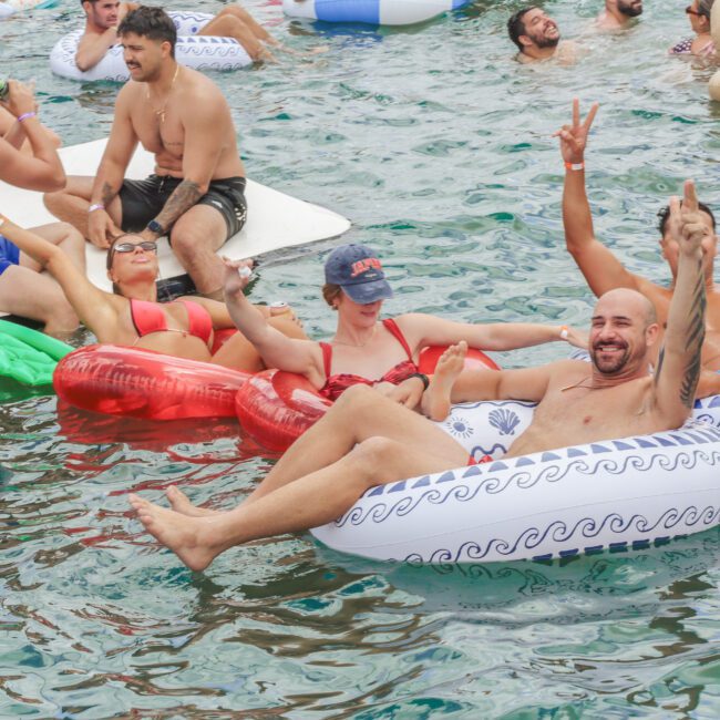 A group of people relaxing on inflatable pool floats in a body of water, smiling and holding drinks. Some are making peace signs, and others are chatting and laughing, enjoying a carefree, sunny day.
