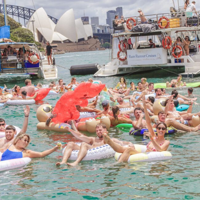 A large group of people on colorful pool floats relax and wave in the water near boats, with the Sydney Opera House and Sydney Harbour Bridge visible in the background under a sunny sky.