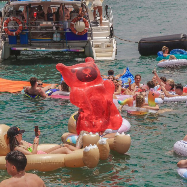 A group of people relax on colorful inflatable rafts and floats, including a large red gummy bear and a brown bear, in the water near a boat, enjoying a lively summer pool party.
