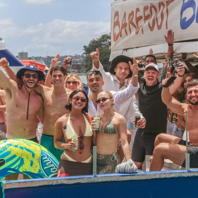 A group of people in swimwear and summer clothing cheer and pose for a photo on a boat during a sunny day, some holding drinks. There is a float and a "Barefoot Blue" sign in the background.