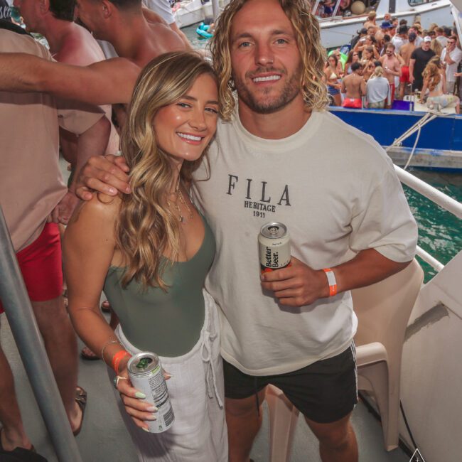 A smiling couple poses together on a crowded boat under sunny skies. The woman wears a green top and white pants while the man is in a white Fila t-shirt and shorts. Both hold drinks, with other people and a boat visible in the background.