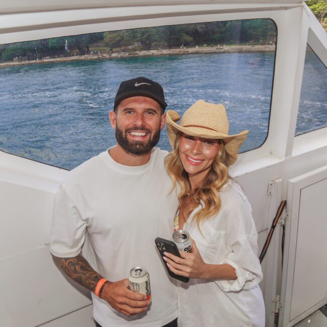 A man and woman smiling and posing together on a boat. The man wears a white T-shirt and black cap, and the woman wears a straw hat and white shirt. Both hold canned drinks, with water and trees visible outside the boat window.
