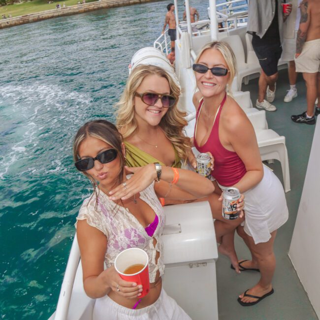 Three women in sunglasses smile and pose on a boat, each holding a drink, with blue water and people in swimwear visible in the background. It’s a sunny day and the mood is festive.