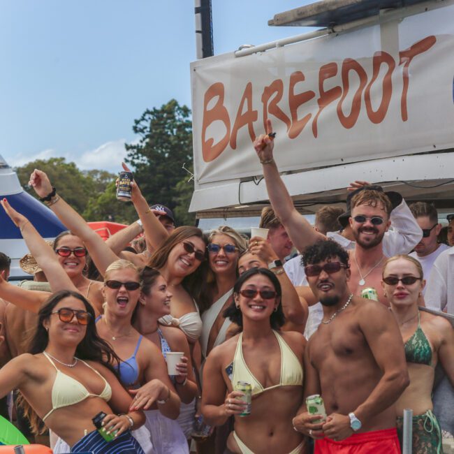 A group of young adults in swimsuits and sunglasses smile and pose with drinks at a lively outdoor boat party under a "BAREFOOT BL" sign, with a boat and blue sky in the background.