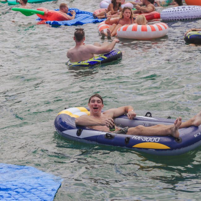 A man relaxes on an inflatable raft in a crowded lake, smiling with his arms and legs outstretched. Other people float nearby on colorful inflatables, enjoying the water.