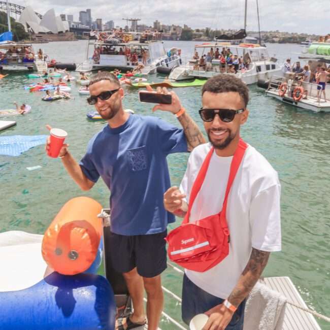 Two men smile and pose with drinks on a boat at a lively party on the water, with many people swimming, relaxing on inflatables, and other boats in the background under a sunny sky.