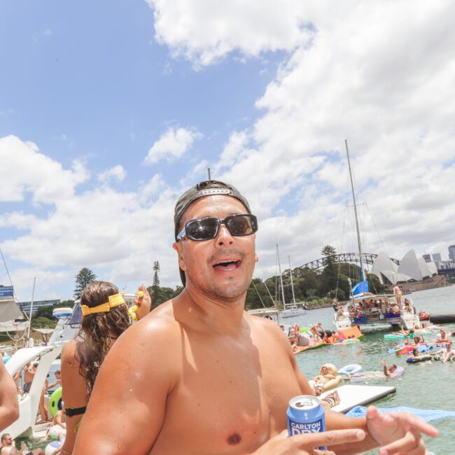 A shirtless man wearing sunglasses and a cap smiles at an outdoor boat party, holding a can of drink. People are swimming or relaxing on boats and inflatables in the water, with city buildings and a bridge in the background.