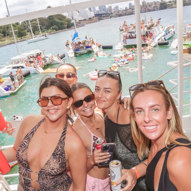 A group of four women in swimsuits and sunglasses smile and pose for a photo aboard a boat; behind them, people enjoy a lively boat party on the water with inflatables and yachts nearby.
