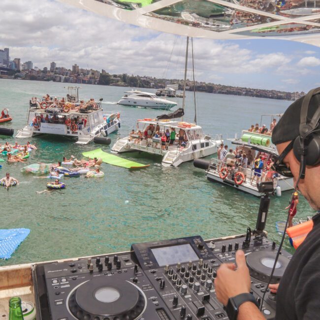 A DJ plays music on a yacht while people relax on nearby boats, floats, and paddleboards in a sunny harbor, with a city skyline and bridge in the background.