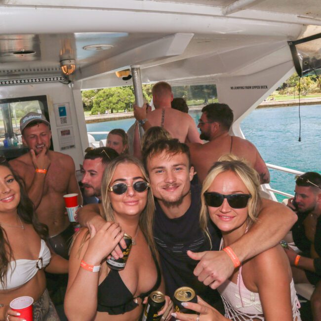 A group of young adults smile and pose together on a boat party. Some hold drinks and wear sunglasses. The boat is crowded, with people socializing in the background and tropical water visible outside.