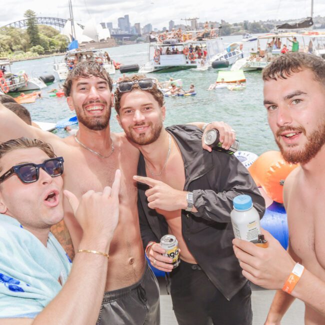 Four men in swimwear smile and pose together at a lively boat party on the water, holding drinks. Other boats, people, and the Sydney Harbour Bridge are visible in the sunny background.