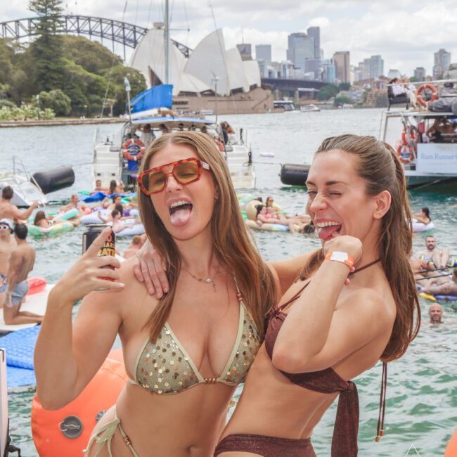 Two women in swimsuits pose and smile at a lively pool party on the water, with boats, people swimming, and the Sydney Opera House and Harbour Bridge visible in the background.