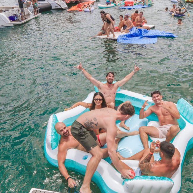 A group of young adults relax and smile on a large inflatable raft in the water, surrounded by other people swimming and lounging on inflatables near boats on a sunny day.