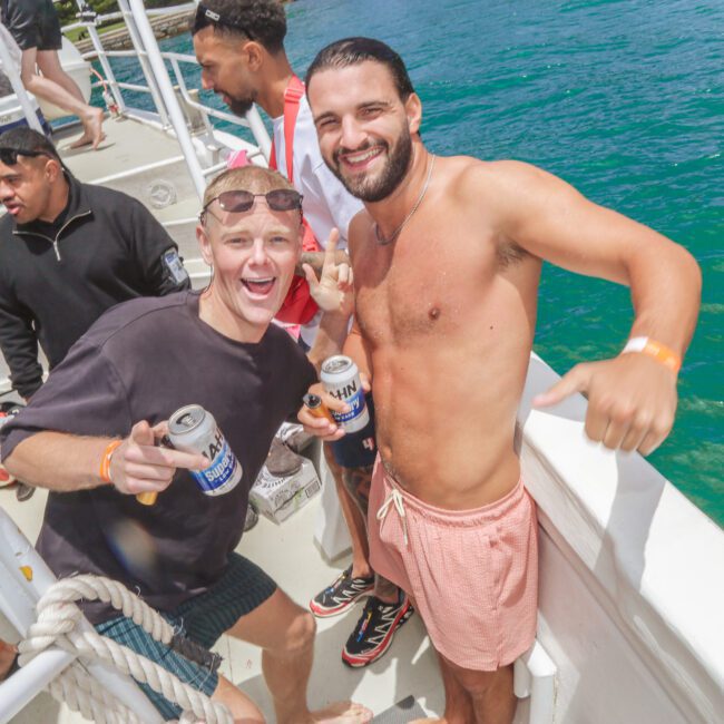 Two smiling men, one shirtless and the other wearing sunglasses, hold cans of beer and pose on a boat with turquoise water in the background. People in swimsuits and casual clothes stand around them, enjoying a sunny day.