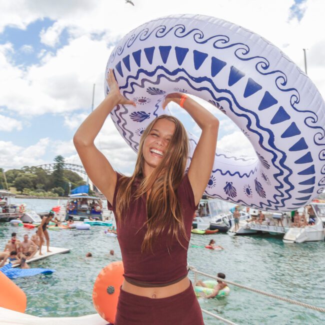A smiling woman stands on a floating dock, holding a large inflatable swim ring above her head. Behind her, people relax on floats and boats on a sunny day on the water.