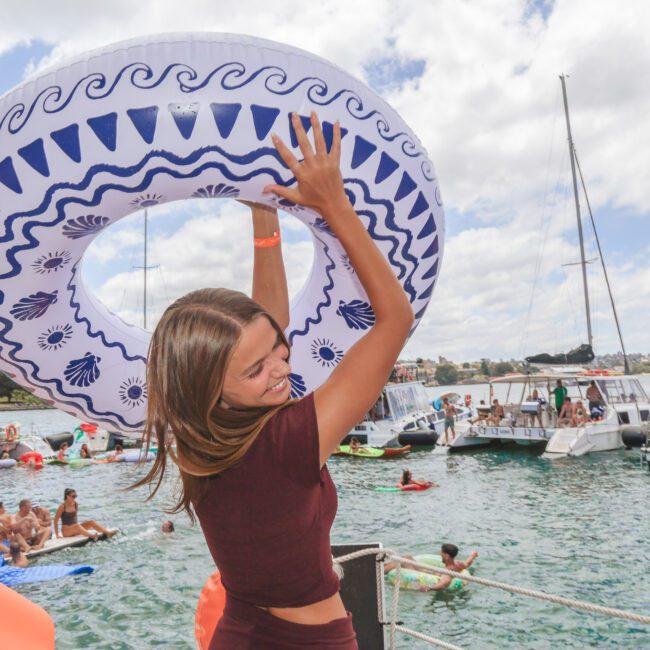 A young woman in a maroon outfit lifts a large, blue-and-white inflatable ring above her head on a dock, surrounded by people enjoying a sunny day on boats and in the water.
