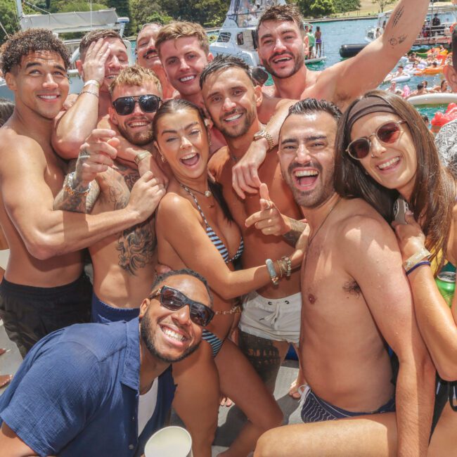 A group of young adults in swimwear smile and pose together on a crowded boat during a sunny day party. Other people and boats are visible in the background on the water.