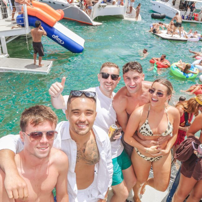 A group of young adults in swimwear smile and pose for a photo at a lively pool party with boats and people on floaties in the background under bright sunny weather.