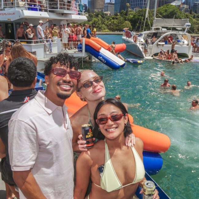 Three people in swimsuits smile at the camera on a boat during a lively summer party, with others swimming and socializing in the water and more boats and city buildings in the background.