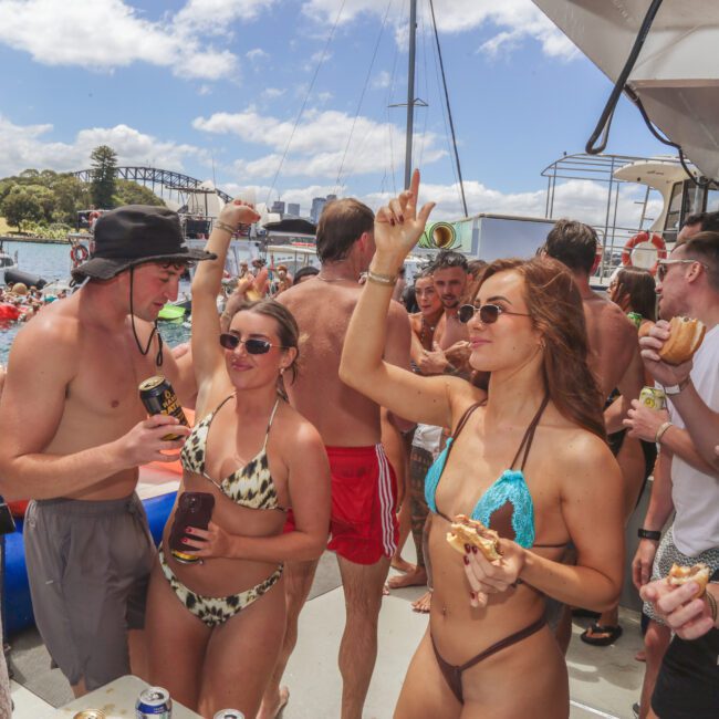 A group of young adults in swimsuits enjoys a lively boat party. Some dance, others eat and drink, with sunny weather and water in the background. People are smiling, wearing sunglasses, and socializing on a crowded deck.