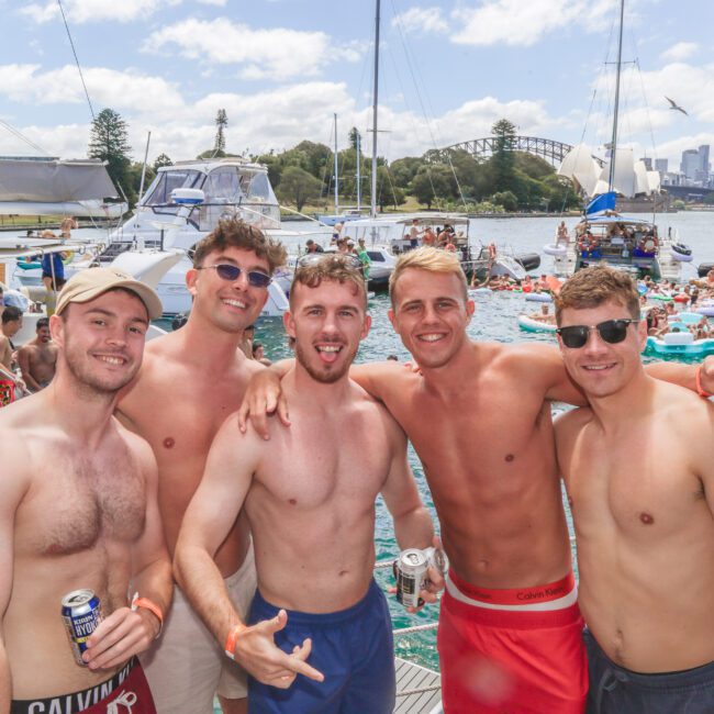 Five young men in swim trunks smile and pose together on a boat at a lively, crowded harbor party, with many people, boats, and inflatables in the background on a sunny day.