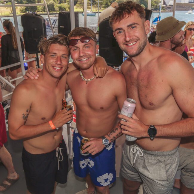 Three smiling men in swim trunks pose together on a boat, holding drinks. Other people in swimwear are around them, enjoying a sunny day on the water. Trees and the river are visible in the background.